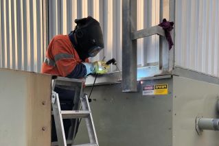 A welder works on metal fabrication indoors, creating bright sparks and smoke.